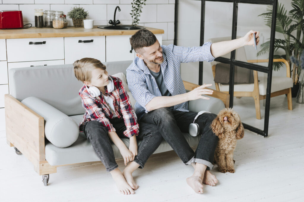 family dad young man and son teenage boy sit on couch in cosy apartment with pet poodle dog and doing selfie on phone, quality time with your family